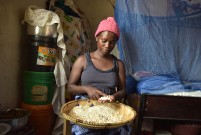 Woman holding a rice bowl and wearing a pink hat and grey shirt sitting in house with blue bednet behind her.